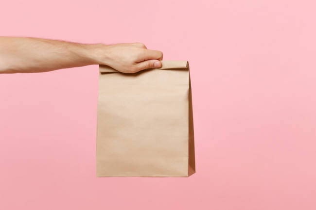Close up male holding in hand brown clear empty blank craft paper bag for takeaway isolated on pastel pink background. Packaging template mock up. Delivery service concept. Copy space advertising area cubitos-de-hielo-no-se-peguen
